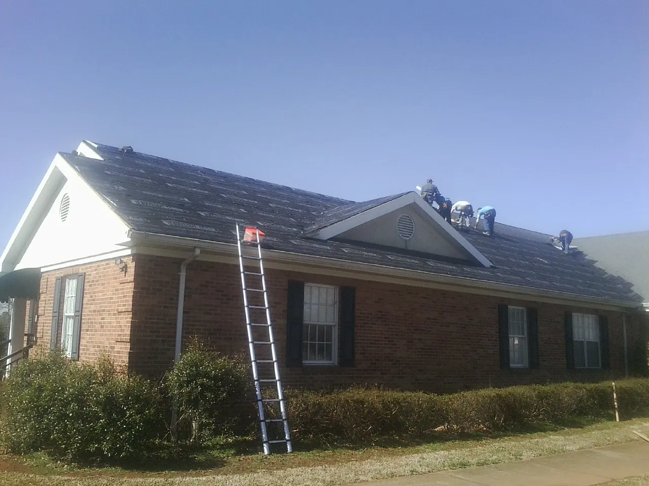 Crew tearing shingles off a brick ranch roof with a ladder set — Emergency Roof Repair work by our crew in Conover